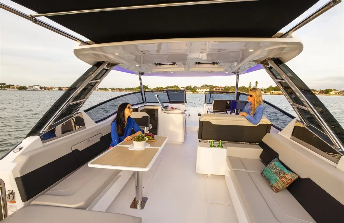 Two women relax on a stylish boat with a table and snacks, overlooking a calm waterway and sunny shoreline.